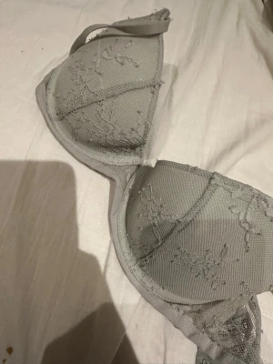 Grågrön spetsdetaljerad BH från H&M - Snygg grågrön BH från H&M med tunna axelband och vacker spetsdetalj på kuporna. Materialet är mjukt och har en lätt glans, vilket ger en lyxig känsla. Perfekt för dig som gillar stilrena underkläder med en touch av spets.