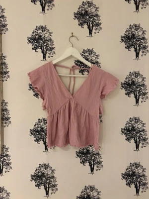 Rosa blus med volang från lager 157 - Säljer en ljusrosa blus från lager 157 i storlek XS. Blusen har v-ringning, korta volangärmar och knytband i ryggen. Materialet är mjukt och krinklat, vilket ger en avslappnad känsla. Perfekt för vår och sommar, och den har en feminin och söt look.