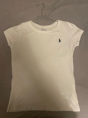 Vit t-shirt från Polo Ralph Lauren - Vit t-shirt från Polo Ralph Lauren med klassisk rund hals och korta ärmar. T-shirten har en liten broderad logga på bröstet och är gjord i mjuk bomull. Enkel och stilren design som funkar till allt, perfekt för dig som gillar clean look.