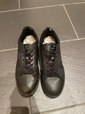 Svarta sneakers från GANT - Säljer ett par svarta sneakers från GANT med klassisk design och rund tå. Skorna har snörning och är tillverkade i skinn med svart platt sula. GANT-logga finns på sidan och bak på hälen. Perfekta för dig som gillar stilrena och enkla sneakers.