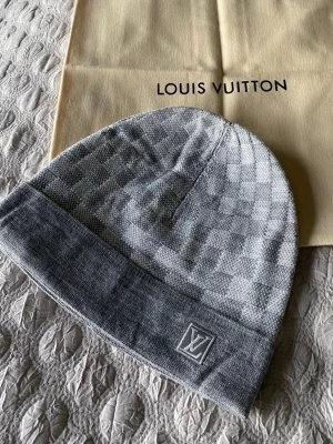 Grå LV Mössa - Snygg grå mössa från Louis Vuitton med klassiskt rutigt mönster och LV-logga framtill. Mössan är stickad och har en bred uppvikt kant för extra stilpoäng. Perfekt accessoar för att lyfta din look under kyliga dagar.