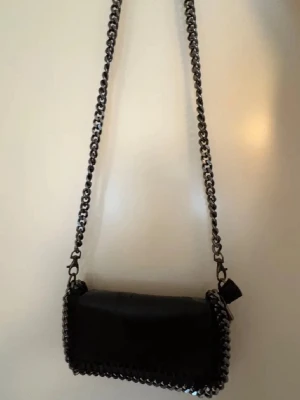 Svart axelväska med kedja - Snygg svart axelväska med grov kedja som axelrem och dekorativ kedjedetalj runt kanterna. Väskan har en rektangulär form och är tillverkad i skinnliknande material. Perfekt för att ge din outfit en cool touch.