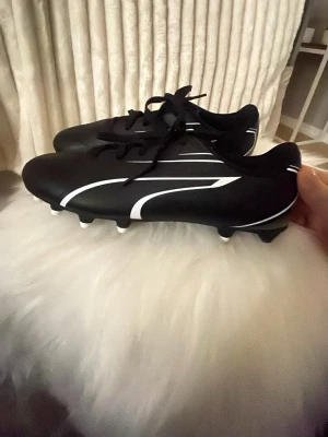Puma Vitoria FG/AG Jr fotbollsskor+puma benskydd  - Svarta fotbollsskor från Puma med vita detaljer och snörning. Modellen Vitoria FG/AG Jr har en rund tå och platt sula med vita dobbar för bra grepp på både gräs och konstgräs. Snygg design med sportiga linjer längs sidorna.Puma benskydd storlek S.Perfekt för dig som älskar fotboll.