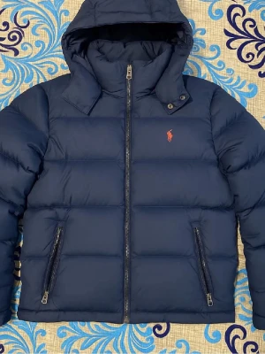 Mörkblå pufferjacka från Polo Ralph Lauren - Säljer en mörkblå pufferjacka från Polo Ralph Lauren med huva och två dragkedjeförsedda fickor. Jackan har det klassiska Polo-logot broderat på bröstet och är tillverkad i polyester. Perfekt för kalla dagar och har en clean, sportig look.