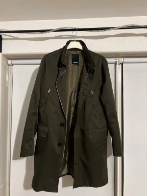 Mörkgrön rock/trenchcoat från J.Lindeberg - Snygg mörkgrön rock/trenchcoat från J.Lindeberg med klassisk krage, två sidofickor med dragkedja och knappar framtill. Jackan har ett bälte med knappar bak och en diskret logga på ärmen. Materialet känns slitstarkt och har en clean look.