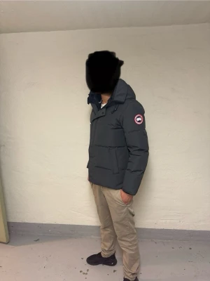 Svart pufferjacka från Canada Goose - Svart pufferjacka från Canada Goose med ikonisk logga på ärmen. Jackan har huva, dragkedja och två stora fickor framtill. Perfekt för kalla dagar med sin tjocka och quiltade design. Stilsäker look och riktigt skön passform. Använd 1-3 gånger för att ta bild med. (Kontakta vid minsta bekymmer)