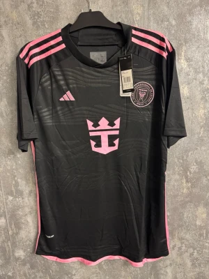 Inter Miami Messi - Ny XL - Säljer en svart Inter Miami matchtröja med rosa detaljer från Adidas, med Messi och nummer 10 på ryggen. Tröjan har rosa ränder på axlarna, klubbmärke och sponsorlogga framtill samt texten 'Freedom to Dream' i nacken. Perfekt för fotbollsfans!