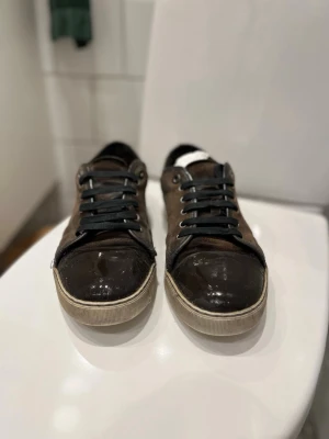 Lanvins - Säljer ett par bruna Lanvins med svart lackad tå och mörkblåa snören. Det är slitna men är snygga ändå. Hälen är lite söndrig men det går enkelt att fixa.