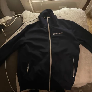 Palm Angels marinblå vindjacka/zip hoodie - Palm Angels vindjacka i marinblått med vita ränder längs ärmarna och vit dragkedja. Jackan har hög krage, två fickor med dragkedja och logotyp på bröstet. Perfekt för dig som gillar streetwear och sportig stil.