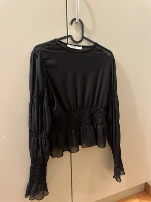 Svart blus med volang i mesh - Säljer en svart blus från NA-KD i storlek XS. Blusen har långa ärmar med smock och volangdetaljer vid ärmslut och midja. Materialet är lätt och transparent mesh som ger en luftig känsla. Perfekt för dig som gillar romantiska detaljer och stilren look. 