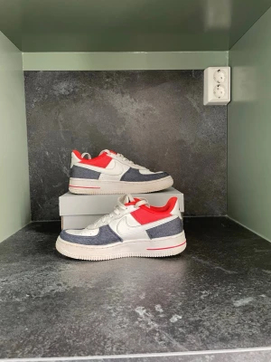 Nike Air Force 1 med denim och röd detalj - Säljer ett par Nike Air Force 1 sneakers med vit läder, denimblå paneler och starkt röda detaljer vid kragen. Skorna har klassisk snörning, rund tå och platt sula. Perfekt för dig som gillar en unik och färgstark stil.
