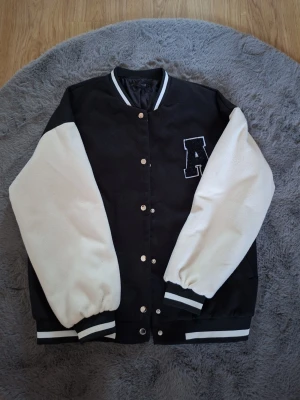 Svart och vit bomberjacka från DAZY - Snygg bomberjacka i svart med vita ärmar och broderad A på bröstet. Jackan har ribbade muddar med vita ränder, tryckknappar framtill och klassisk college-look. Perfekt för dig som vill ha en cool och sportig stil.
