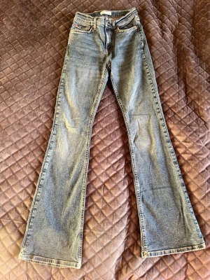 Bootcut jeans från Gina Tricot, strl 34 - Bootcut jeans från Gina Tricot i klassisk blå tvätt. Modellen heter Perfect Jeans och har fem fickor, bälteshällor och dragkedja med knapp. Jeansen är tillverkade i jeansmaterial och har en snygg bootcut-passform som ger en retro vibe. Det finns en liten slitning på baksidan. Högt upp. Se bilder. Går att sy igen med några stygn. 