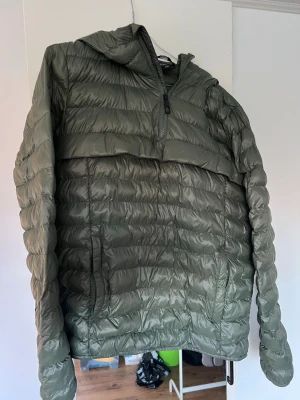 Grön pufferjacka från Everest - Säljer en grön pufferjacka från Everest med huva och halv dragkedja. Jackan har ett quiltat mönster och är tillverkad i syntetmaterial. En liten liten vit fläck, se foto, annars i fint skick.