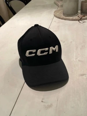 Svart Flexfit keps från CCM - Säljer en svart keps från CCM med Flexfit-passform. Kepsen har vit broderad CCM-logga både fram och bak, samt ventilerande meshpaneler med små hål för extra komfort. Perfekt för dig som gillar sportig stil och vill ha en snygg accessoar. Finns knappt att köpa längre pga att det är den äldre modellen