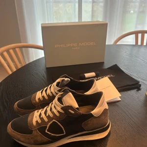 Philippe Model grå/blå sneakers - Snygga Philippe Model sneakers i grått och marinblått med vita detaljer och snörning. Skorna har en rund tå, platt sula och är tillverkade i mocka och textil. Märkets logga syns på sidan och bak på hälen. Perfekta för dig som gillar stilrena och sportiga sneakers. Dem är också perfekta nu till vintern. Dem är också väldigt sköna att gå i. Skorna är i storlek 40 men är lite stora i storlek. Dem är i bra skick men har börjat tappa lite i bak sulan som går att fixa lätt