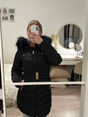 Svart vinterjacka med bälte - Säljer en svart vinterjacka med snyggt bälte i midjan och dragkedja framtill. Jackan har huva med svart fuskpäls och gulddetaljer på dragkedjor och snören. Bältet sitter inte fast i jackan men följer med