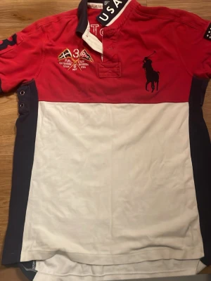 Röd, vit och marinblå Polo Ralph Lauren skjorta - Säljer en snygg Polo Ralph Lauren chief keef skjorta i rött, vitt och marinblått med broderade detaljer, stor USA-flagga på ärmen och stort tryck med siffran 3 på ryggen. Skjortan har korta ärmar, klassisk krage och knappar framtill. Perfekt för dig som gillar sportig och amerikansk stil. Sitter som S 