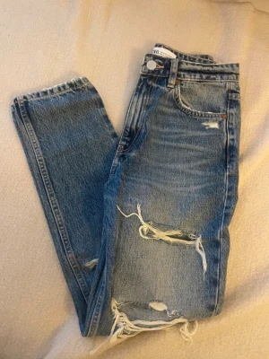 Blå ripped jeans från Zara, strl 34 - Säljer ett par blå jeans från Zara i storlek 34 med hög midja och raka ben. Jeansen har flera stora slitningar och hål framtill för en riktigt edgy look. Klassisk femficksmodell i kraftigt bomullstyg. Perfekta för dig som gillar streetstyle och vill sticka ut.