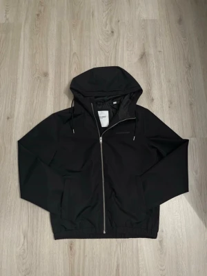 Svart vindjacka från Jack & Jones - Svart vindjacka från Jack & Jones med huva och snörning. Jackan har dragkedja framtill, två sidofickor och elastisk nederkant. Materialet är syntet och jackan har en stilren look med diskret logga på bröstet. Perfekt för blåsiga dagar.