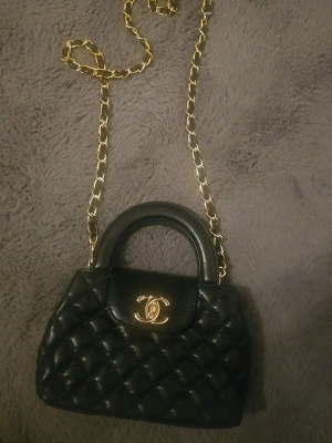 Svart quilted väska - Säljer en svart quiltad handväska i ikonisk guldfärgad CC-logga på locket. Väskan har både handtag och en snygg kedja i guld för axelbärande. Tillverkad i skinn med elegant rutmönster och praktiskt fack inuti. Perfekt för dig som gillar lyxiga detaljer och klassisk stil.