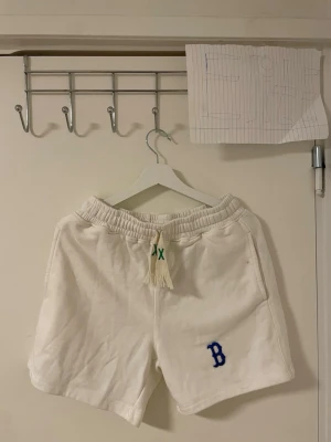 Vita MLB shorts med blå B,  - Säljer ett par vita shorts från MLB i storlek S. De har en broderad blå B på framsidan, grön snörning i midjan och en bakficka. Mjukt material och elastisk midja gör dem riktigt sköna. Perfekta för chill dagar och sportiga outfits.Liknande modell som originala.