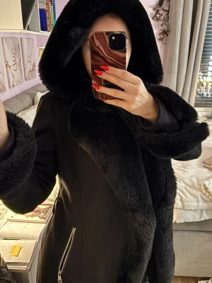 Svart pälsjacka med stor huva - Säljer en svart pälsjacka med stor fluffig huva och breda pälsdetaljer längs krage och ärmslut. Jackan har dragkedja i sidan och är riktigt mysig och varm. Dock saknas bältet.