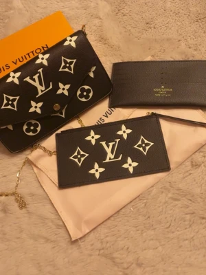 Louis Vuitton svart kuvertväska med kedja - Svart kuvertväska från Louis Vuitton med ikoniskt monogrammönster i vitt. Väskan har guldfärgad kedja och detaljer, samt matchande plånbok och korthållare. Tillverkad i skinn med tryckknapp framtill och snyggt LV-emblem. Perfekt för dig som gillar lyxiga accessoarer.