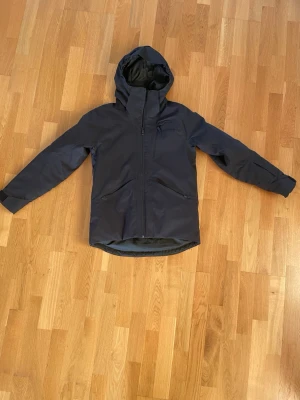 Svart vindjacka från Everest med huva - Svart vindjacka från Everest med snygg huva och dragkedja framtill. Jackan har två praktiska fickor med dragkedja och en diskret logga på bröstet. Materialet är slitstark polyester och passformen är normal. Perfekt för blåsiga dagar och outdoor.