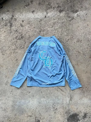 Blå långärmad mesh-topp Chrome Hearts - Säljer en blå långärmad mesh-topp från Chrome Hearts med stora tryck i vitt och turkos på ryggen och ärmarna. Toppen har perforerat material och coola grafiska detaljer med texten 'HEARTS' och 'CH'. Perfekt för dig som gillar streetwear och unika plagg.