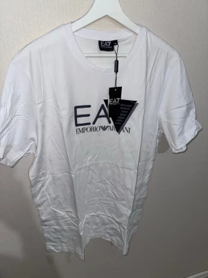 Vit EA7 Emporio Armani t-shirt XXL - Säljer en fräsch vit t-shirt från EA7 Emporio Armani i storlek XXL. T-shirten har korta ärmar och ett stort tryck med EA7-loggan på bröstet. Materialet är mjuk bomull som känns skönt mot huden. Perfekt för dig som gillar stilrena och exklusiva märken.T-shirten kan också passa till storlek XL. Hör av dig via frågor⁉️