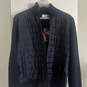 hybridjacka från J.Lindeberg - Hybridjacka i mörkblått från J.Lindeberg med quiltad front och stickade ärmar. Jackan har dragkedja, ståkrage och två sidofickor. Primaloft-material i fronten för extra värme och stilren look. Perfekt för dig som gillar sportig och modern design.