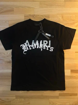 Svart AMIRI Brothers t-shirt - Svart t-shirt från AMIRI med stor vit 'Brothers'-text i gotisk stil över bröstet och AMIRI-logga bakom. Baksidan har vit text med budskap och små grafiska symboler. Klassisk rund hals och korta ärmar. Materialet är mjuk bomull, perfekt för en chill och stilren look.