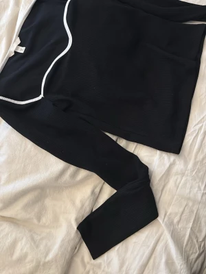 Svart långärmad topp från H&M - Svart långärmad topp från H&M med vit ribbad kant längs halsringningen. Toppen har en snygg vågig detalj vid halsen och är gjord i ett ribbat material som ger en cool struktur. Perfekt att styla med jeans eller kjol.