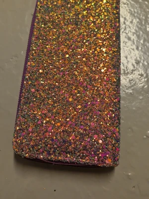 Glittrig plånbok med lila dragkedja - Säljer en plånbok med glittrig framsida i orange, rosa och guld. Plånboken har lila dragkedja och flera fack för kort och mynt. Perfekt för dig som gillar färg och glitter! Materialet är syntet och insidan är svart. Aldrig använt