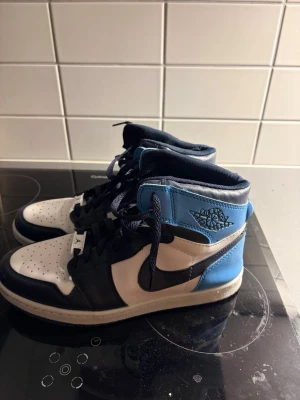 Nike Air Jordan 1 Mid blå och vit - Säljer ett par Nike Air Jordan 1 Mid sneakers i blått, vitt och marinblått skinn. Klassisk high-top siluett med perforerad tå, svart swoosh och snörning. Ikonisk Jordan Wings-logga på sidan och mjuk textil på insidan för extra komfort.