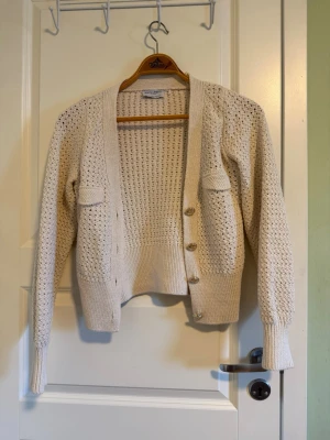 Beige stickad kofta från Holly & White - Säljer en beige stickad kofta från Holly & White med snyggt hålmönster och ribbade muddar. Koftan har långa ärmar, två fickor framtill och knäpps med fyra knappar. Perfekt för lager-på-lager och chill stil.