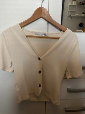 Beige croppad blus med knappar - Säljer en beige croppad blus från 157 med korta ärmar och v-ringad hals. Toppen har tre dekorativa knappar framtill och är gjord i mjukt bomullsmaterial. Perfekt för dig som gillar enkel och stilren design. Har aldrig använt🫰🏼💞 om ni vill ställa några frågor så gör gärna det 