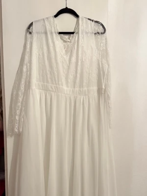 Vit långklänning med spetsdetaljer - Säljer en elegant vit långklänning med spets upptill och långa spetsärmar. Klänningen har v-ringad hals och dragkedja i ryggen. Till klänningen ingår även ett vitt slöja med kam och ett par matchande spetsvantar. Perfekt för speciella tillfällen.