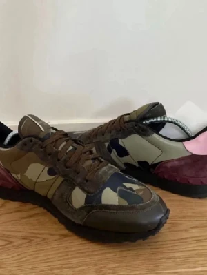 Valentino Rockrunner sneakers camouflage - Säljer ett par Valentino Rockrunner sneakers med camouflagemönster i grönt, brunt och marinblått. Skorna har detaljer i mocka och skinn, samt en cool kontrast med rosa och vinröd häl. Svart platt sula och snörning framtill. Perfekta för dig som gillar statement-sneakers.