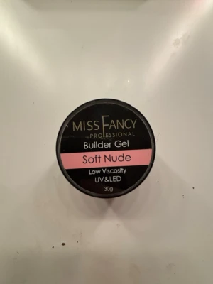 Miss Fancy Builder Gel Soft Nude - Builder Gel Soft Nude från Miss Fancy Professional, 30g. Gelen har en mjuk nude-rosa ton och låg viskositet, perfekt för att bygga och förstärka naglar. Passar både UV och LED-lampa. Kommer i en rund svart burk med rosa etikett.