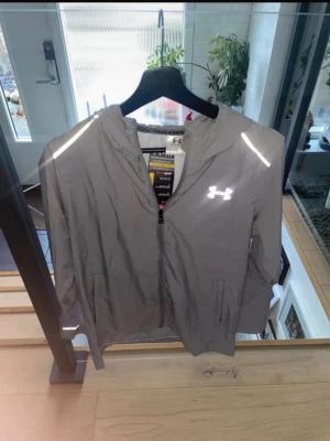 Grå vindjacka från Under Armour - Snygg grå vindjacka från Under Armour med huva och reflekterande detaljer på axlar, ärmslut och logga. Jackan har dragkedja framtill och fickor på sidorna. Perfekt för löprundan eller när du vill ha en sportig look. Ny med prislapp