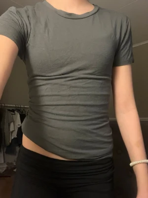 Mörkgrön basic t-shirt - Säljer en enkel och stilren mörkgrå t-shirt med rund hals och korta ärmar. Modellen är tight och passar perfekt till jeans eller shorts. Materialet känns mjukt och skönt mot huden, troligtvis bomull. Perfekt för dig som gillar minimalistisk stil.