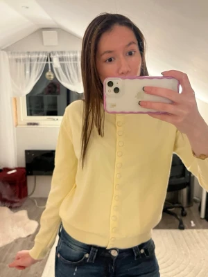 Gul kofta från ONE-K med knappar - Söt ljusgul kofta från ONE-K med rund hals och matchande tygklädda knappar hela vägen fram. Mjuk och skön, perfekt till vår och sommar. Långärmad och i ett tunt, stickat material som känns fräscht och stilrent. Obs står ingen storlek men passar som s!