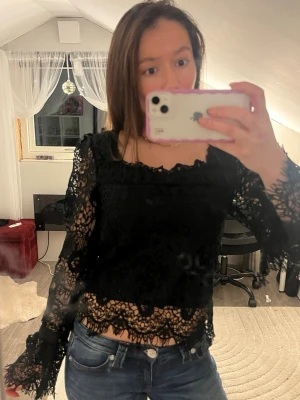 Svart spetsblus med öppen rygg - Säljer en svart blus i spets med långa ärmar och fina detaljer. Toppen har öppen rygg med knytband och dragkedja, samt vackra volangkanter. Perfekt för dig som gillar unika och snygga toppar med mycket detaljer. Obs det står ingen storlek men passar som xs-s!