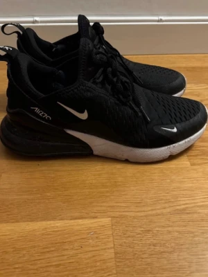 Nike Air 270 svarta sneakers - Svarta Nike Air 270 sneakers med vit swoosh och detaljer. Ovandel i mesh för bra andningsförmåga, snörning och en tydlig Air-enhet i hälen. Sulan är vit med röda och svarta partier undertill. Perfekta för dig som gillar sportig och modern stil.