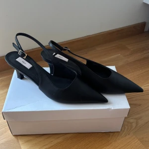 Svarta pumps med spetsig tå från Nelly - Använda ca 2 gånger. Stilrena svarta pumps från Nelly med spetsig tå och låg klack. Skorna har en smal rem med spänne runt hälen och är tillverkade i syntetmaterial. Perfekta för dig som gillar en klassisk och elegant look.