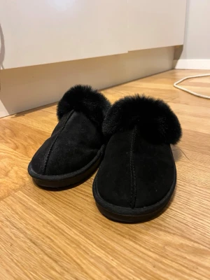 Svarta fluffiga tofflor från 157 - Mysiga svarta tofflor med fluffig päls runt öppningen och mjuk mockaliknande yta. Perfekta att glida runt i hemma när du vill ha det extra skönt om fötterna. Sulan är platt och stabil, och hela skon har en rund tå för maximal komfort.