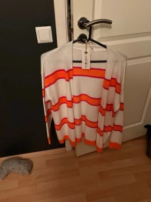 Randig kofta från Key Largo - Säljer en mjuk och skön kofta från Key Largo i vitt med orange och rosa ränder. Koftan har öppen front och långa ärmar, perfekt för att slänga över en t-shirt eller linne. Materialet känns stickat och har en avslappnad passform.