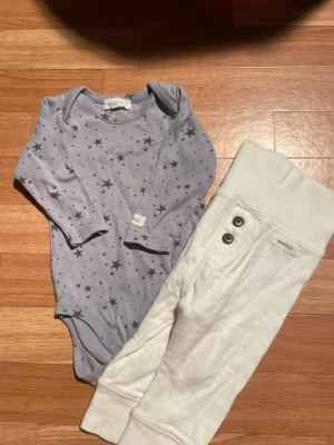 Ingen väst på bilden - Bilden visar en långärmad body med stjärnmönster och ett par vita byxor storlek är 68 märke newbie